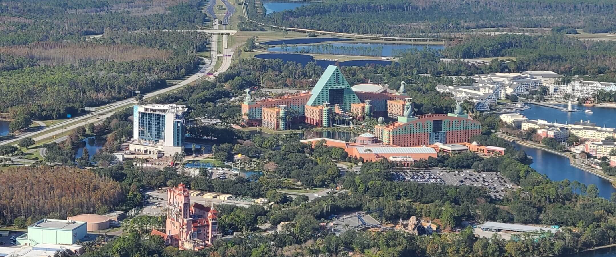 Aerial view of a resort complex with large buildings, including a distinctive triangular-roofed structure, surrounded by trees, roads, lakes, and parking lots. A tall, pink building with spires is also visible in the foreground.