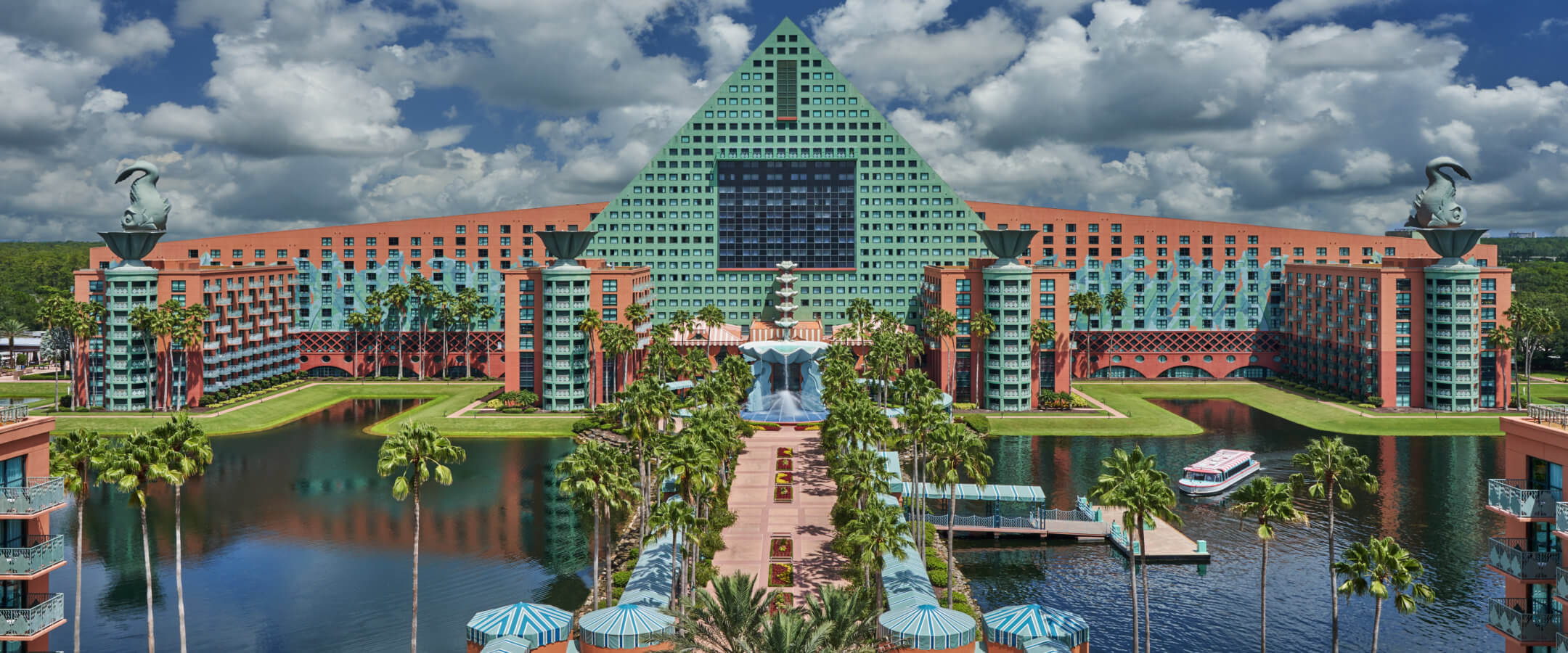 A large, colorful hotel with a green triangular roof and two black swan statues on top, surrounded by water, palm trees, and a blue sky with fluffy clouds.