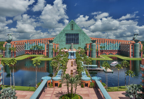 A large, colorful hotel with a green triangular roof and two black swan statues on top, surrounded by water, palm trees, and a blue sky with fluffy clouds.