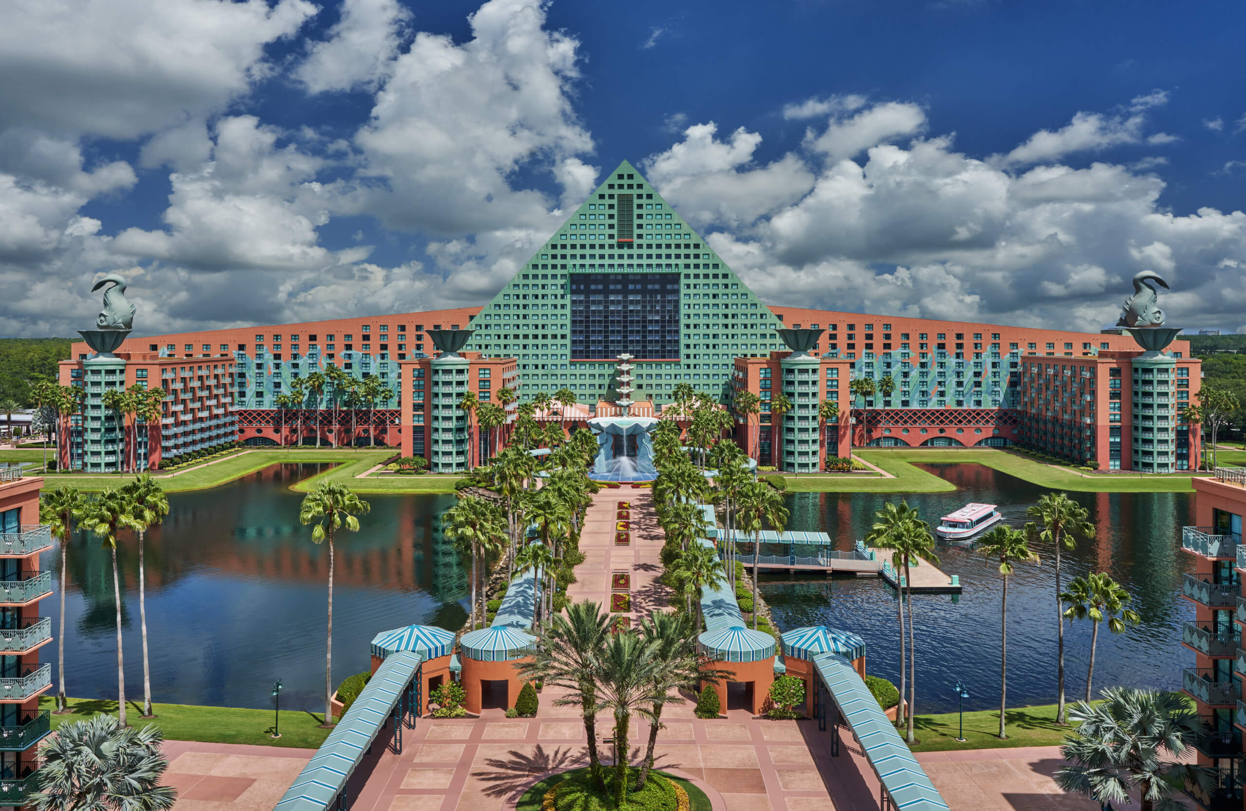 A large, colorful hotel with a green triangular roof and two black swan statues on top, surrounded by water, palm trees, and a blue sky with fluffy clouds.