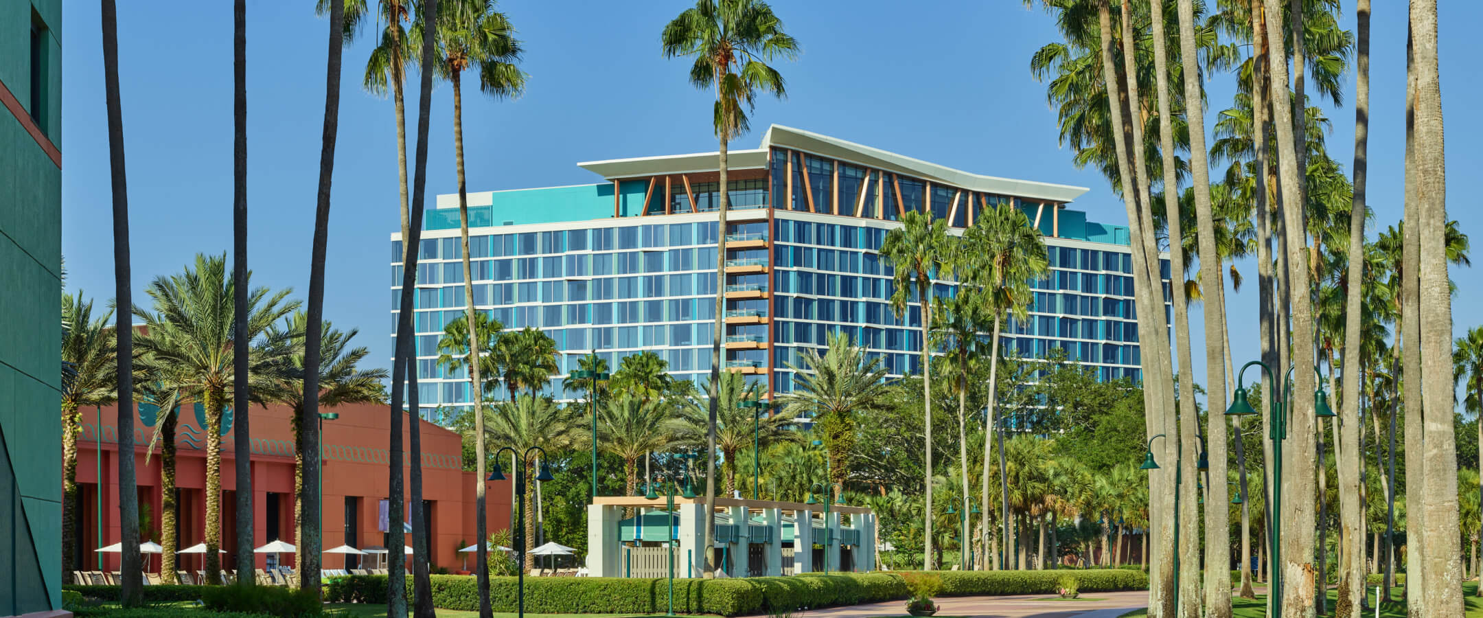 A modern, blue-glass hotel rises behind a landscaped walkway lined with tall palm trees on a sunny day, with clear blue skies and lush greenery surrounding the scene.