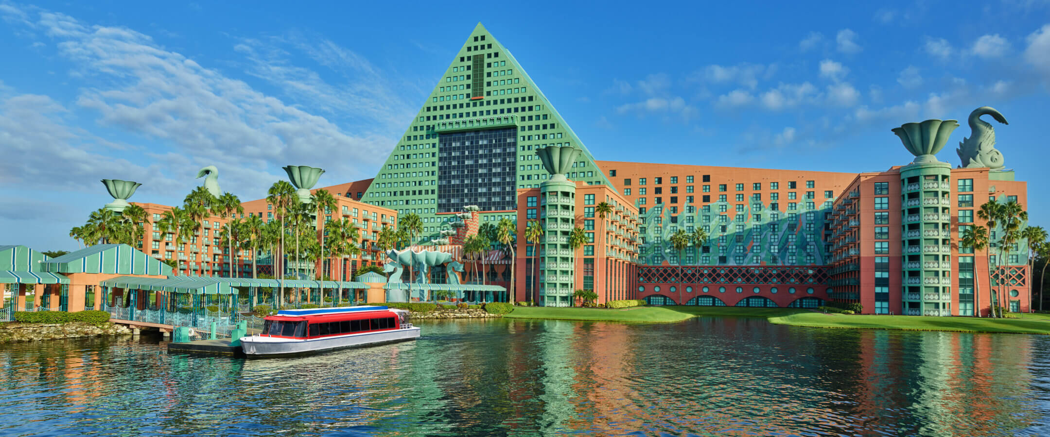A large, colorful hotel with a green triangular roof and unique sculptures sits beside a calm lake, with a red and white boat docked at the waterfront under a bright blue sky.