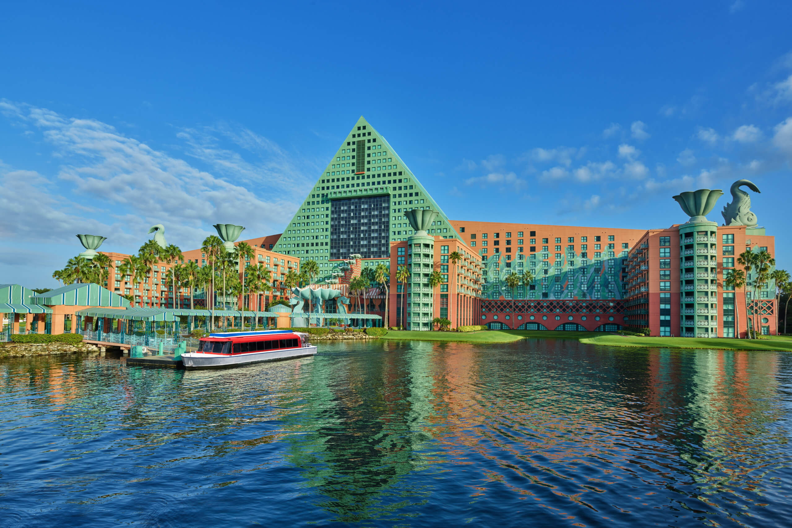 A large, colorful hotel with a green triangular roof and unique sculptures sits beside a calm lake, with a red and white boat docked at the waterfront under a bright blue sky.