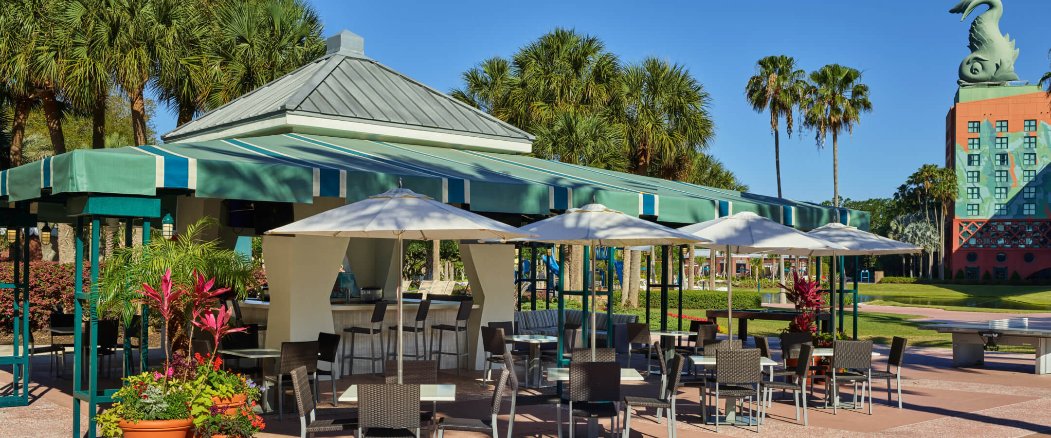 Outdoor patio seating area with tables, chairs, and umbrellas under a green awning, surrounded by palm trees and plants, with a whimsical hotel building visible in the background on a sunny day.