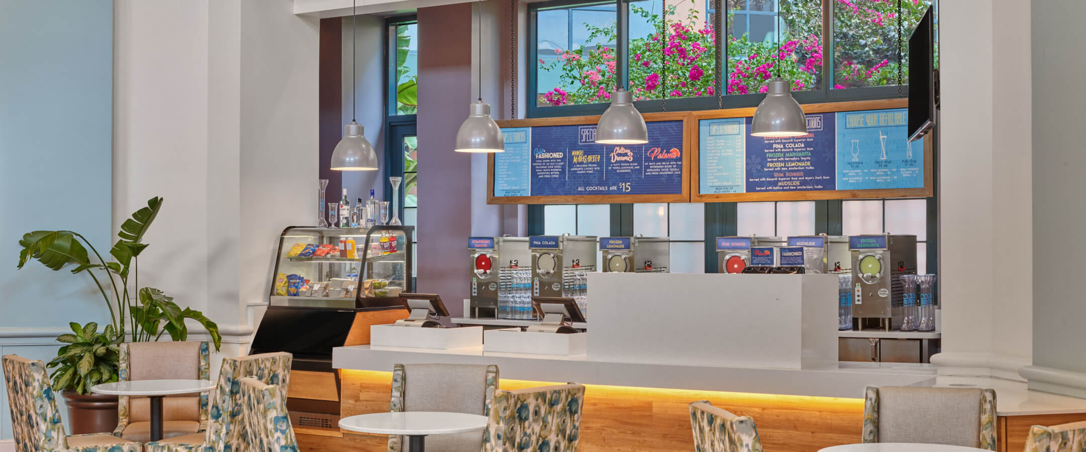 A modern café with patterned chairs and round tables. The counter features a menu board, pastries display, hanging pendant lights, and large windows above with views of flowers outside.