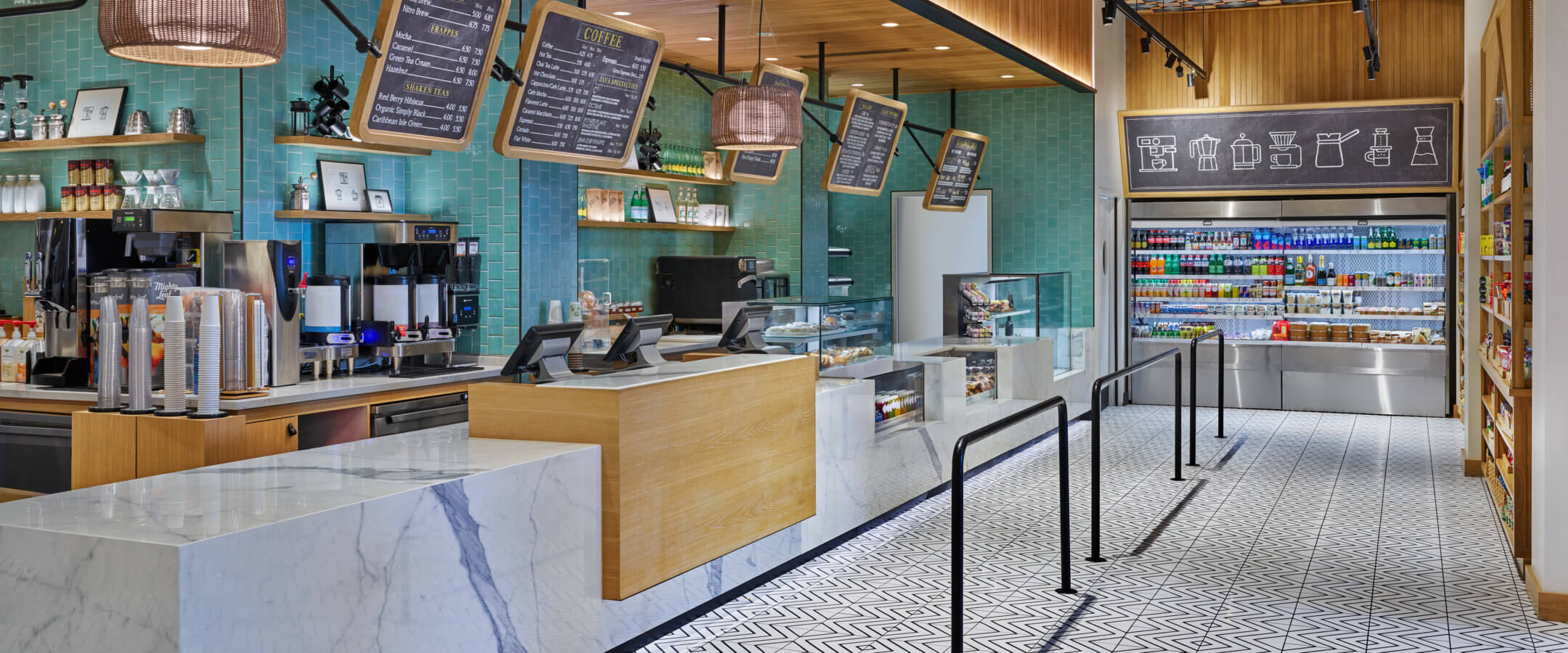 A modern café interior with marble counters, digital menu boards, turquoise tiled walls, geometric floor tiles, beverage refrigerators, pastry displays, and hanging lights, creating a bright and inviting atmosphere.