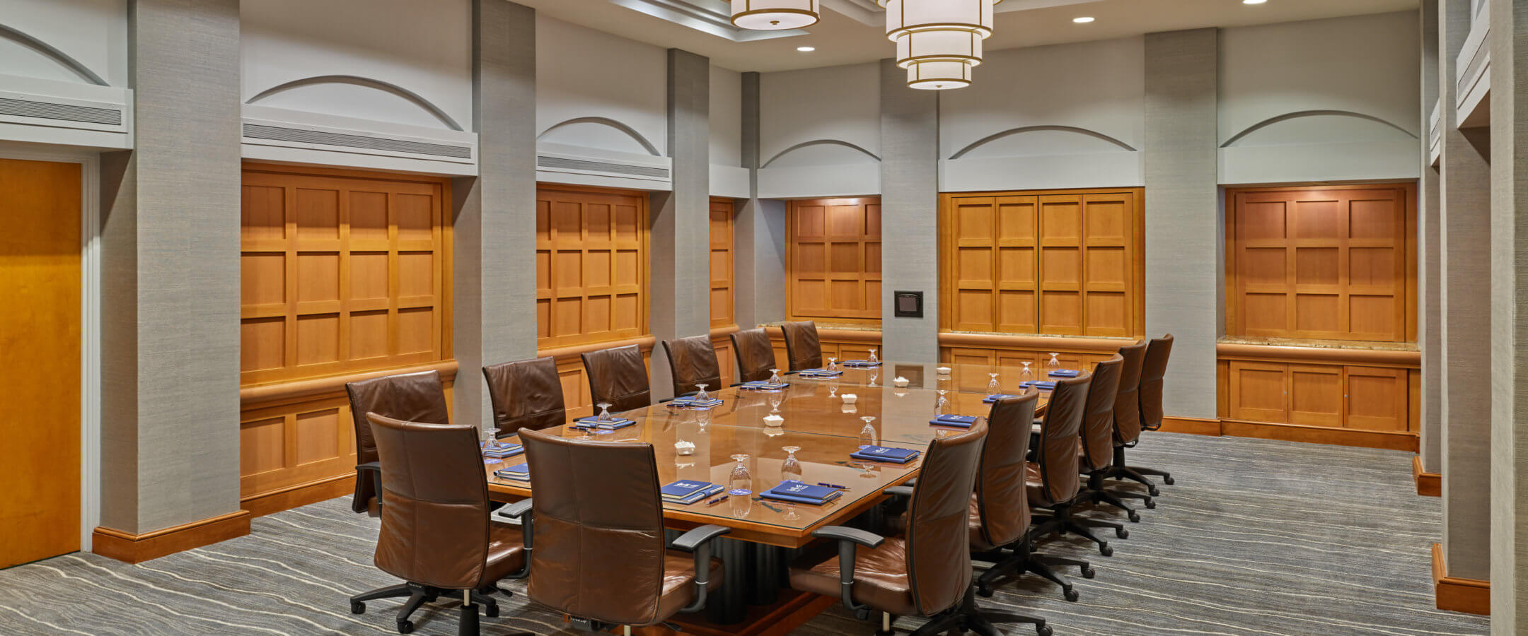 A modern conference room with a large rectangular wooden table surrounded by brown leather chairs, set with glasses, notepads, and napkins, under contemporary ceiling lights.