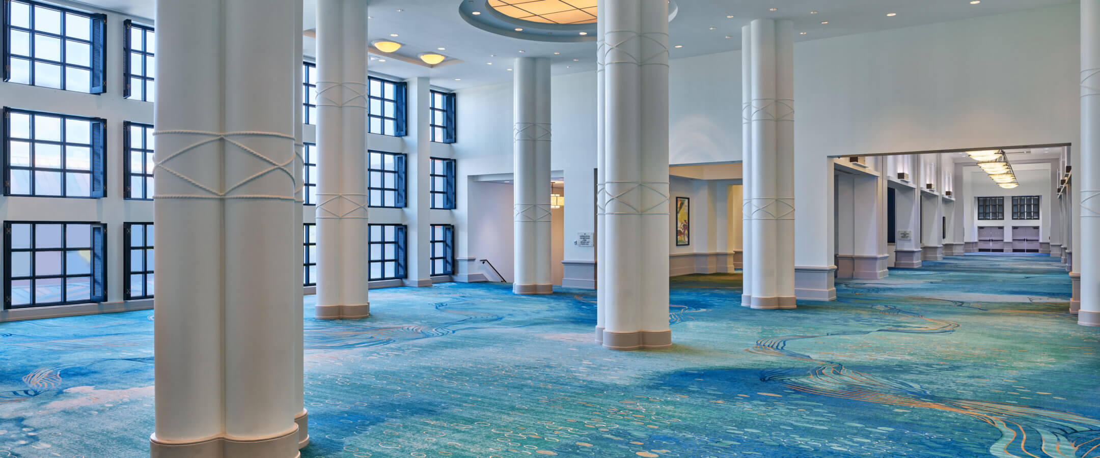 Spacious, elegant hall with tall white columns, large round ceiling lights, blue patterned carpet, and floor-to-ceiling windows letting in natural light.