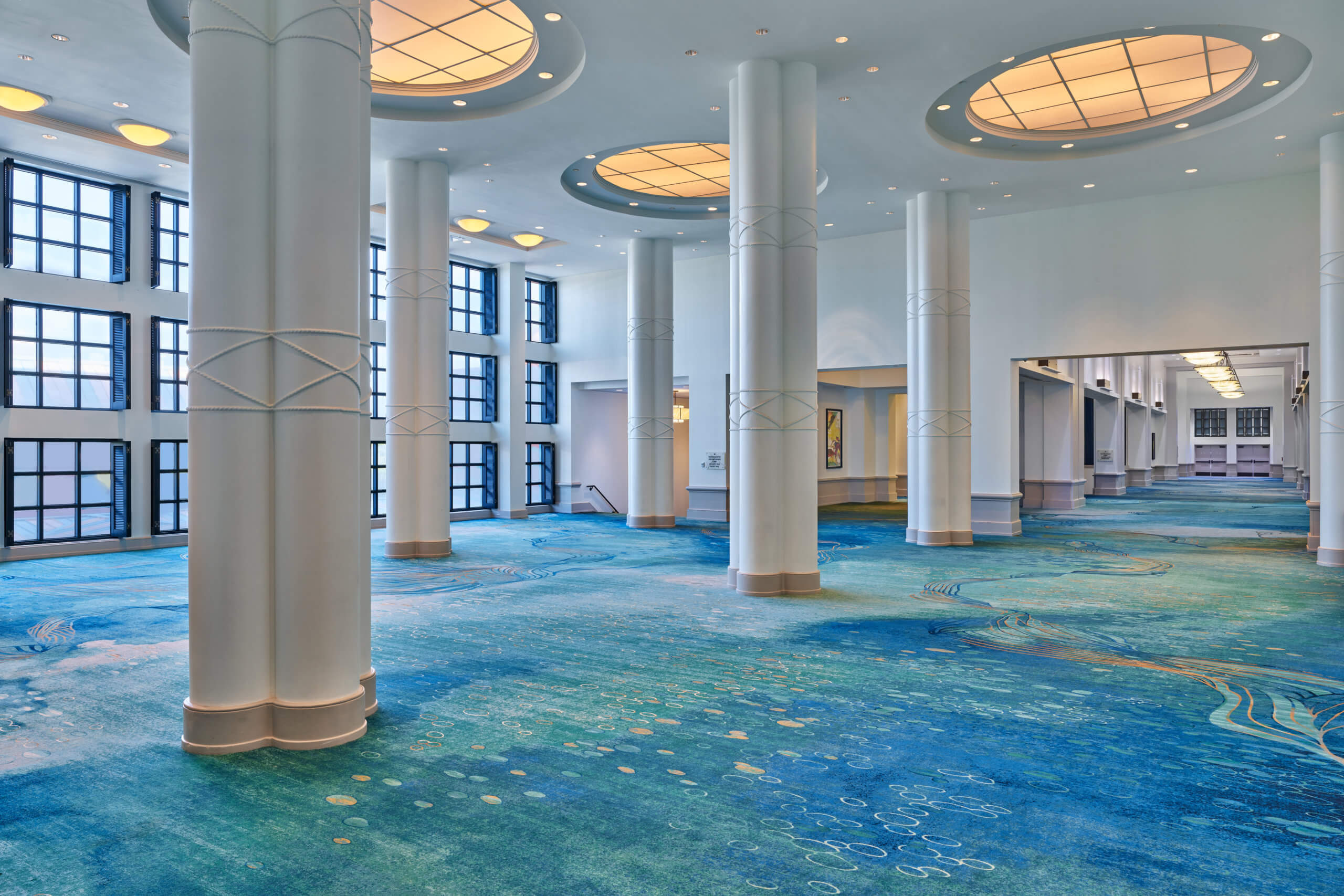 Spacious, elegant hall with tall white columns, large round ceiling lights, blue patterned carpet, and floor-to-ceiling windows letting in natural light.