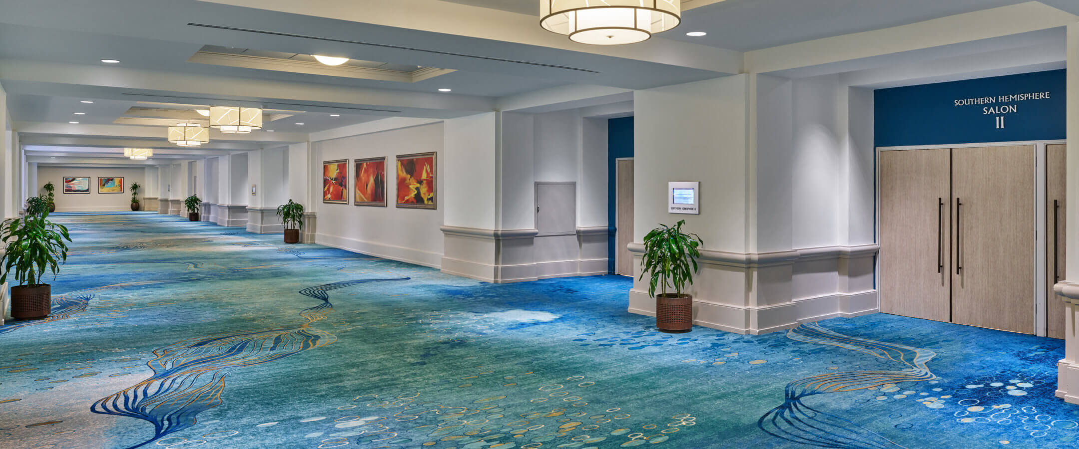 A spacious hotel hallway with blue patterned carpet, white walls, ceiling lights, large potted plants, colorful abstract paintings, and a closed door labeled Southern Hemisphere Salon II.