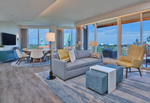 A modern, bright living and dining area with large windows showing a cityscape, gray sofa, blue and yellow accents, dining table with chairs, TV, and wooden floors. Natural light fills the open, airy space.