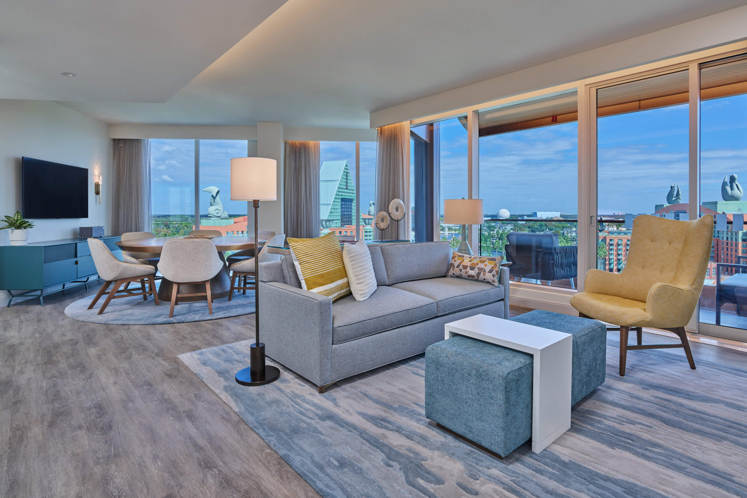 A modern, bright living and dining area with large windows showing a cityscape, gray sofa, blue and yellow accents, dining table with chairs, TV, and wooden floors. Natural light fills the open, airy space.