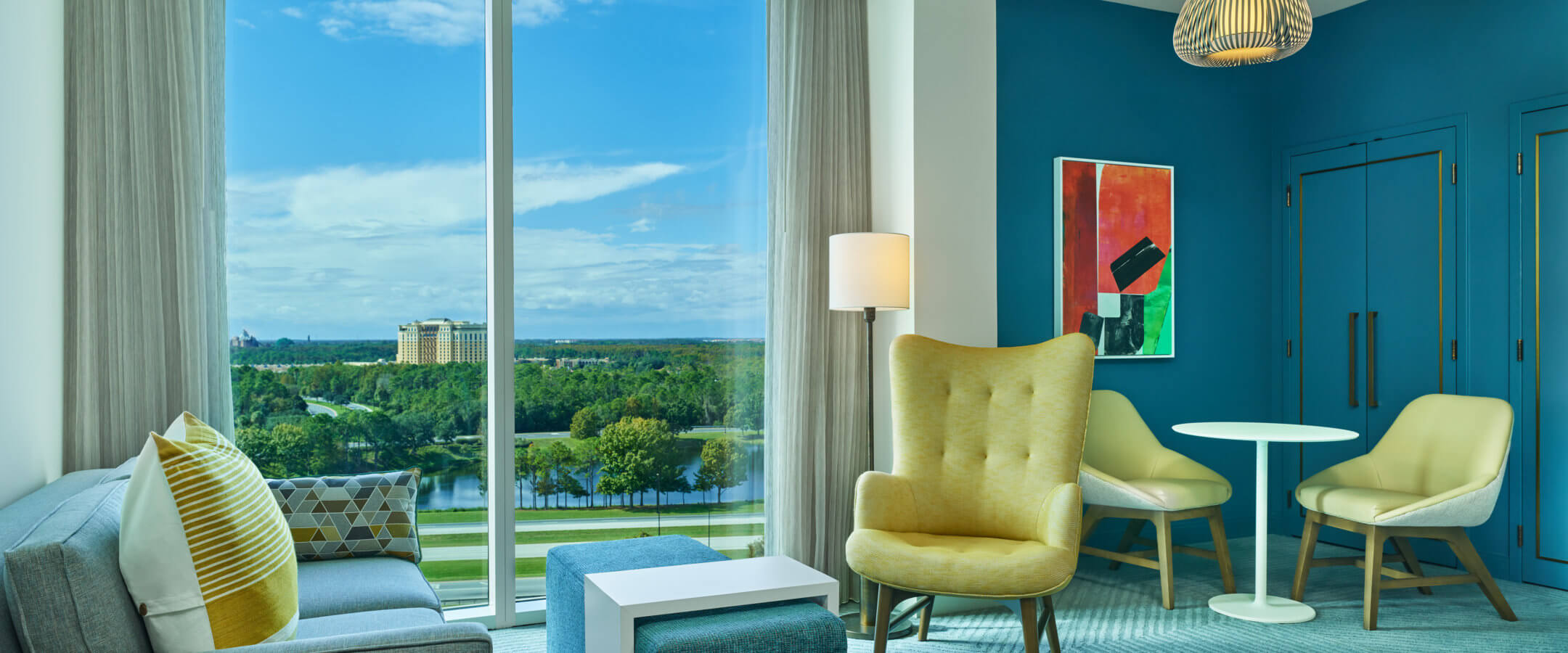 A modern hotel room with large floor-to-ceiling windows showing a scenic view of trees and a building. Inside are stylish chairs, a sofa, a small table, modern decor, and blue accent walls.