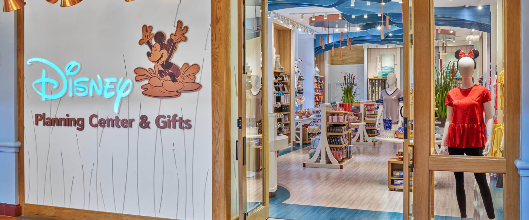 A Disney Planning Center & Gifts store entrance with a neon Disney sign, Mickey Mouse graphic, and a mannequin in a red shirt with Minnie Mouse ears. Inside, shelves display Disney-themed merchandise.