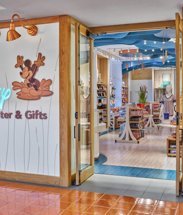 A Disney Planning Center & Gifts store entrance with a neon Disney sign, Mickey Mouse graphic, and a mannequin in a red shirt with Minnie Mouse ears. Inside, shelves display Disney-themed merchandise.