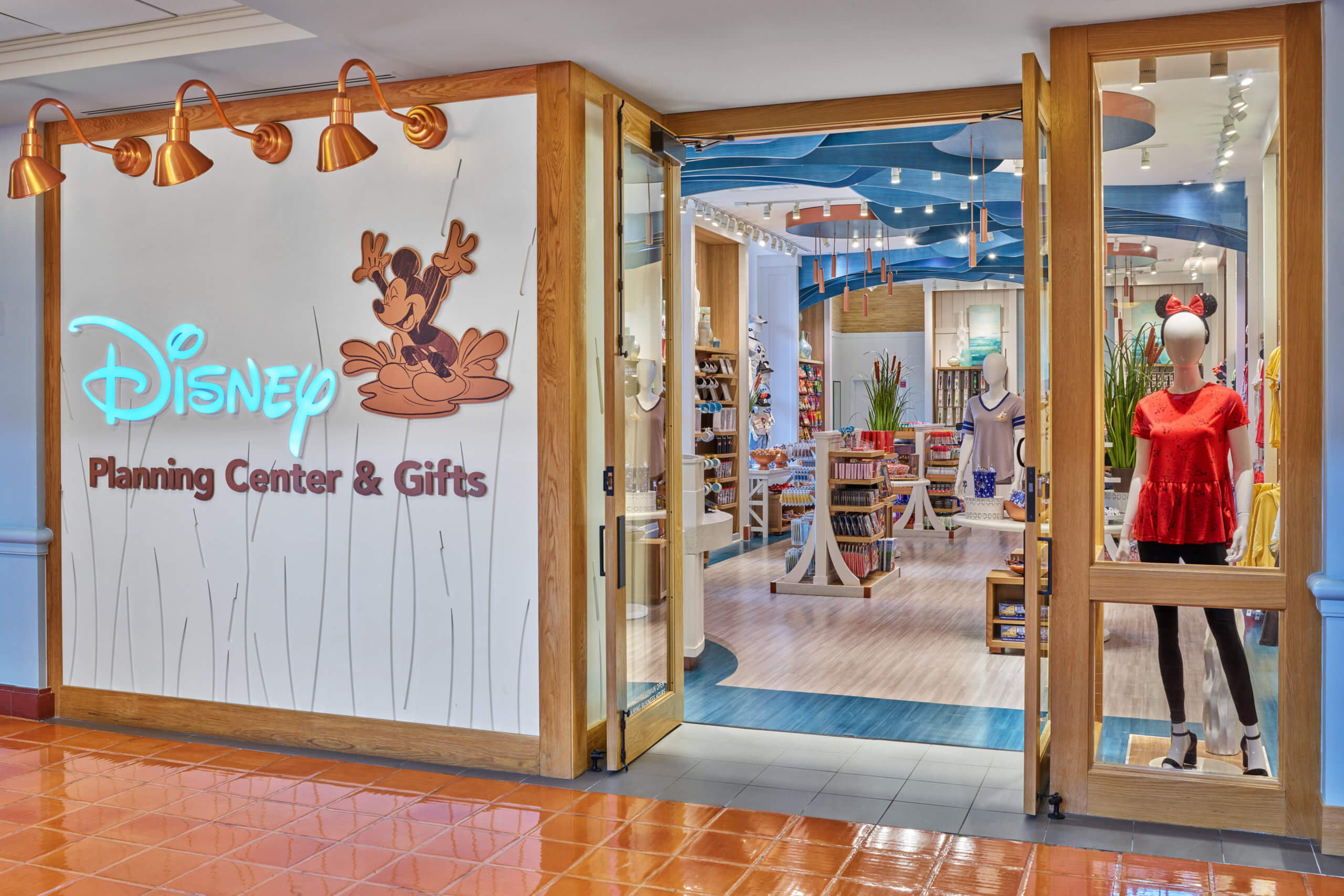 A Disney Planning Center & Gifts store entrance with a neon Disney sign, Mickey Mouse graphic, and a mannequin in a red shirt with Minnie Mouse ears. Inside, shelves display Disney-themed merchandise.