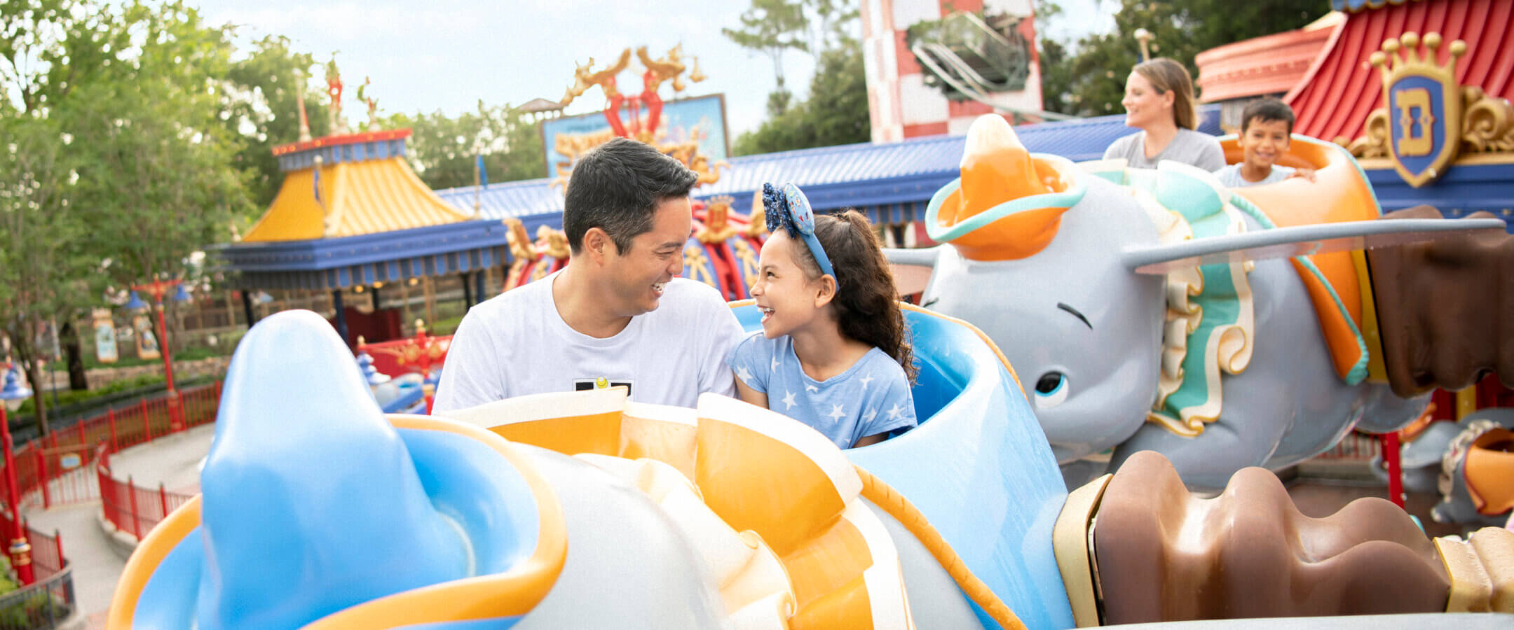 A smiling father and daughter ride a flying Dumbo attraction at an amusement park, surrounded by colorful ride vehicles and cheerful visitors in the background.