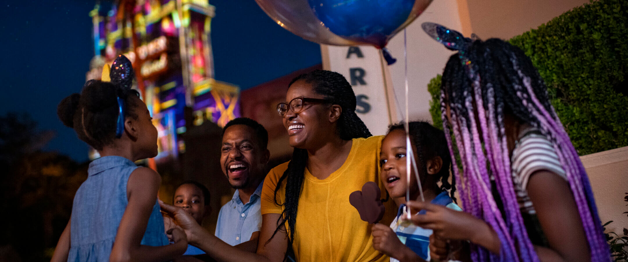 A joyful family of five sits together at night, laughing and celebrating. One child holds a Mickey-shaped balloon, and another holds a Mickey ice cream bar. The family wears festive attire, with two children in Minnie Mouse ears.