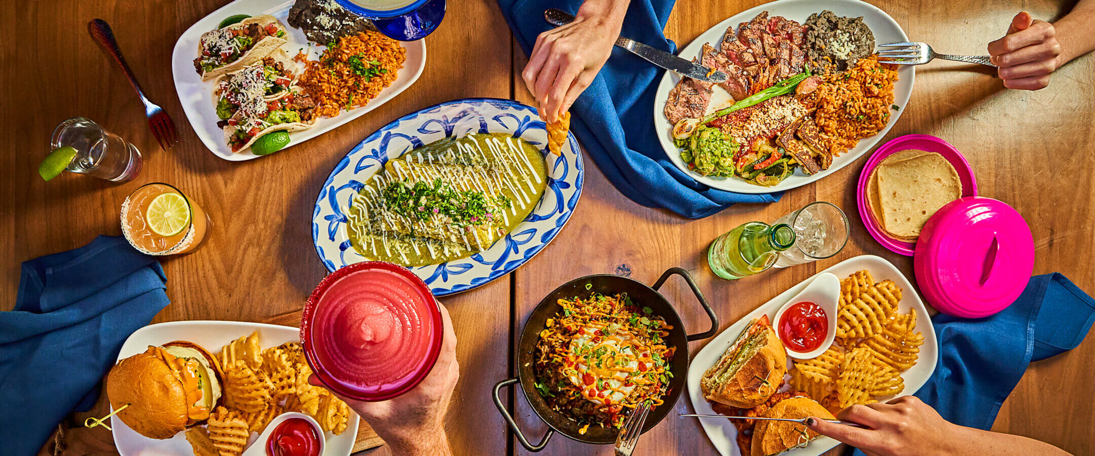 A table set with various Mexican dishes including tacos, enchiladas, rice, beans, fries, a burger, grilled fish, and drinks. Multiple hands reach for food and drinks, creating a lively dining scene.