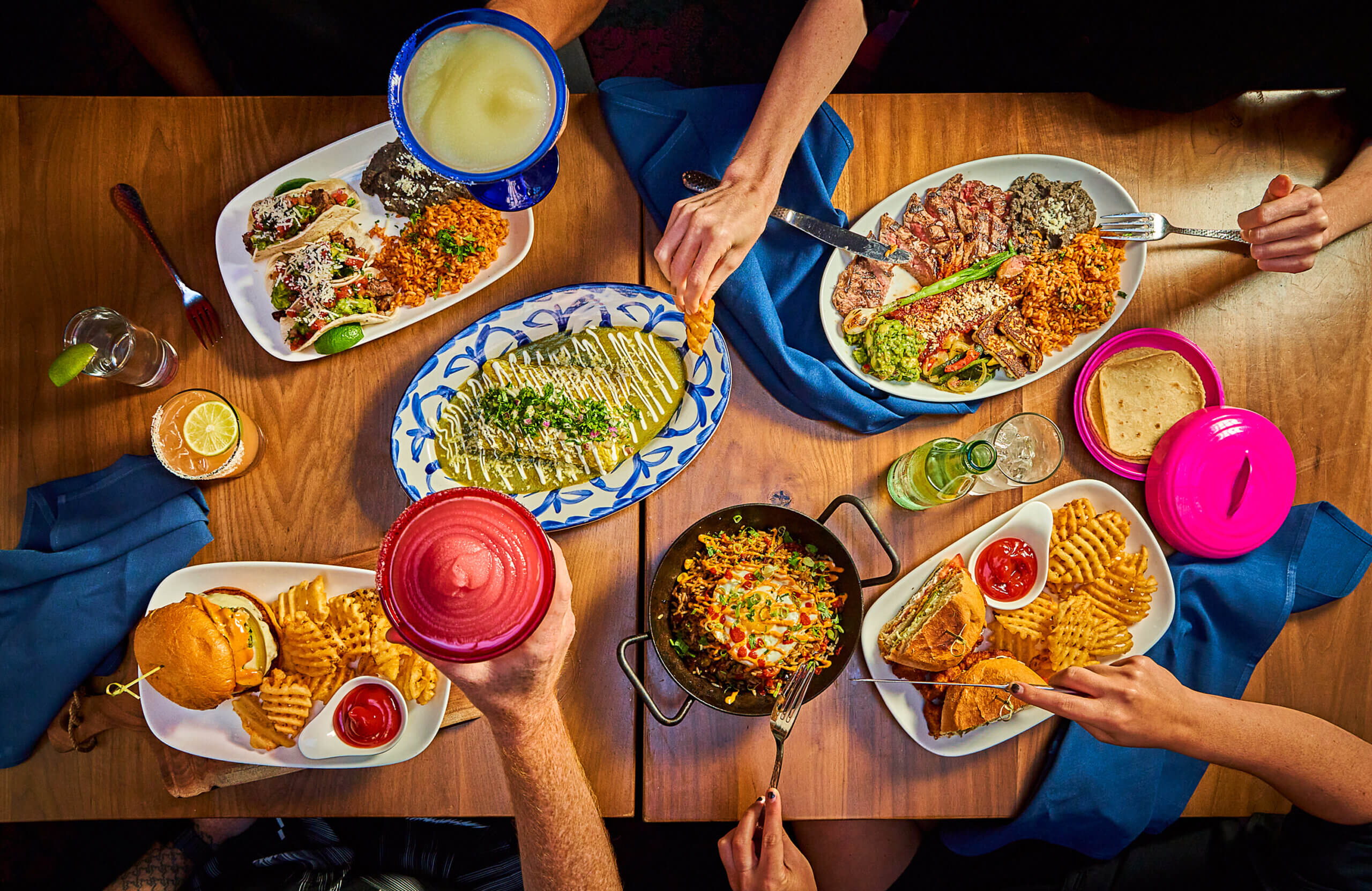Une table garnie de divers plats mexicains, dont des tacos, des enchiladas, du riz, des haricots, des frites, un hamburger, du poisson grillé et des boissons. Plusieurs mains se tendent vers la nourriture et les boissons, créant ainsi une scène de repas animée.
