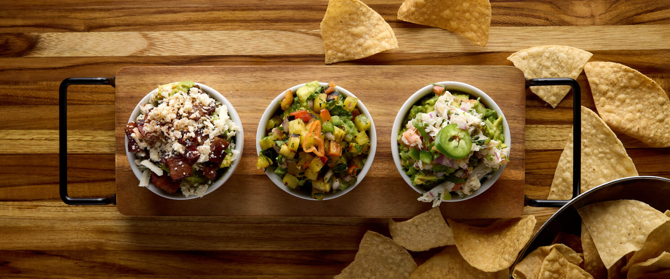 Three small bowls of colorful salsa and dip on a wooden board with handles, surrounded by scattered tortilla chips on a wooden table.