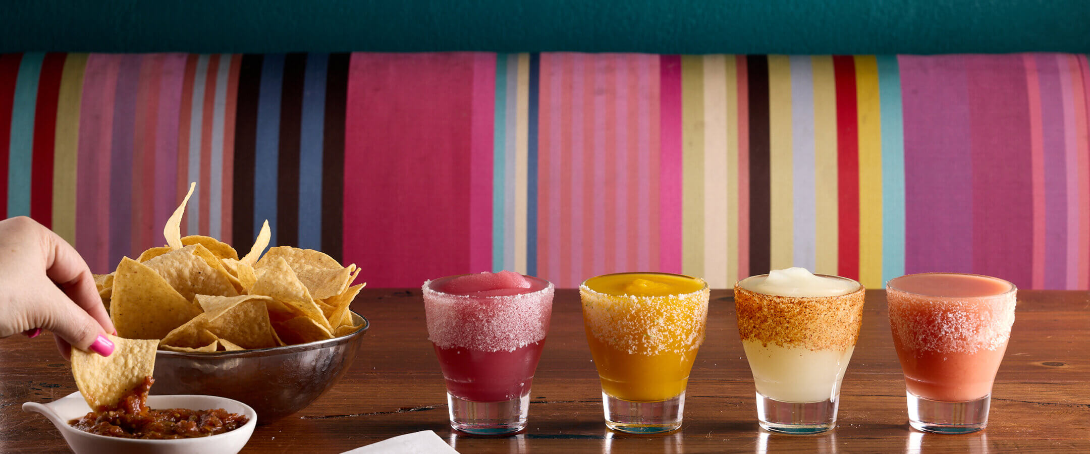 A hand dips a tortilla chip into salsa next to a bowl of chips. In front, five colorful margaritas in salt-rimmed glasses are lined up on a wooden table, with a vibrant striped booth in the background.