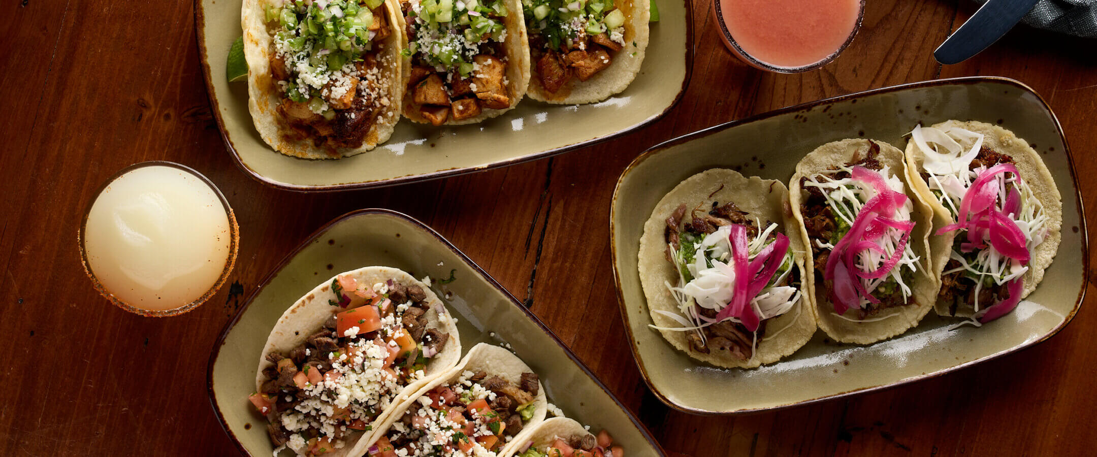 Three plates of assorted tacos with various toppings sit on a wooden table, accompanied by four colorful drinks in round glasses. Each plate holds three tacos with different fillings and garnishes.