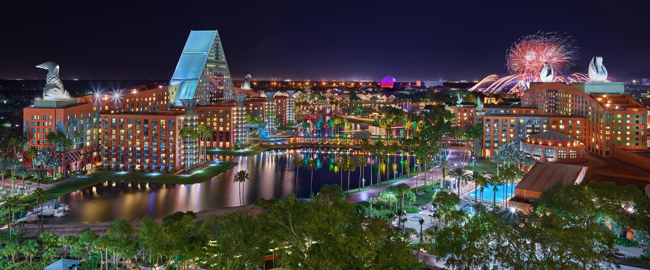 Nighttime view of the illuminated Walt Disney World Swan and Dolphin Resort with fireworks in the background, reflecting on a central lake surrounded by trees and pools.