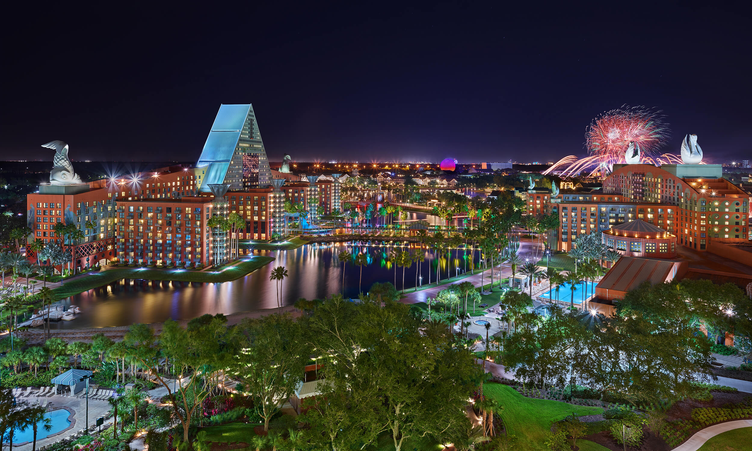 Ночной вид на освещенный Dolphin Resort  Walt Disney World Swan and Dolphin Resort с фейерверками на заднем плане, отражающийся в центральном озере, окруженном деревьями и бассейнами.