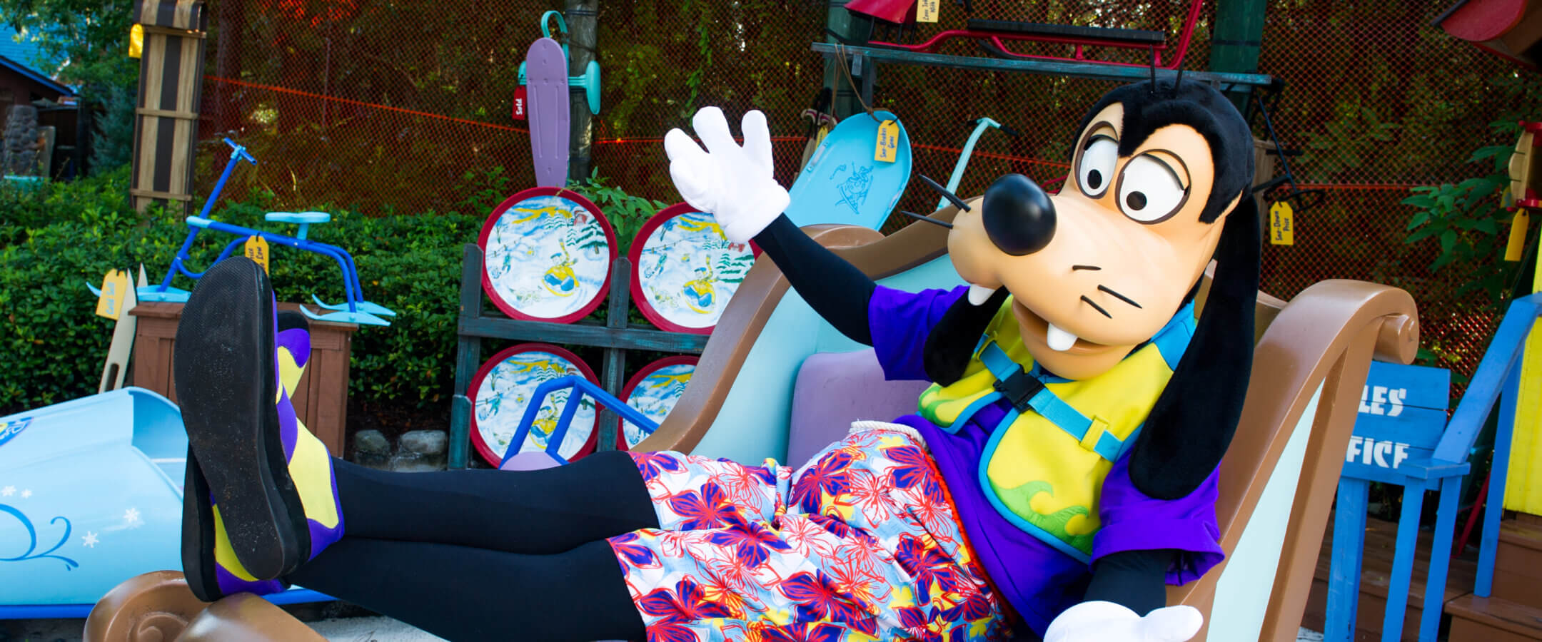 Goofy, wearing colorful shorts and a purple shirt, sits casually on a sleigh with his feet up, surrounded by snow-themed decorations and signs like Low Mile Sleds and Buy Now, Sleigh Later.