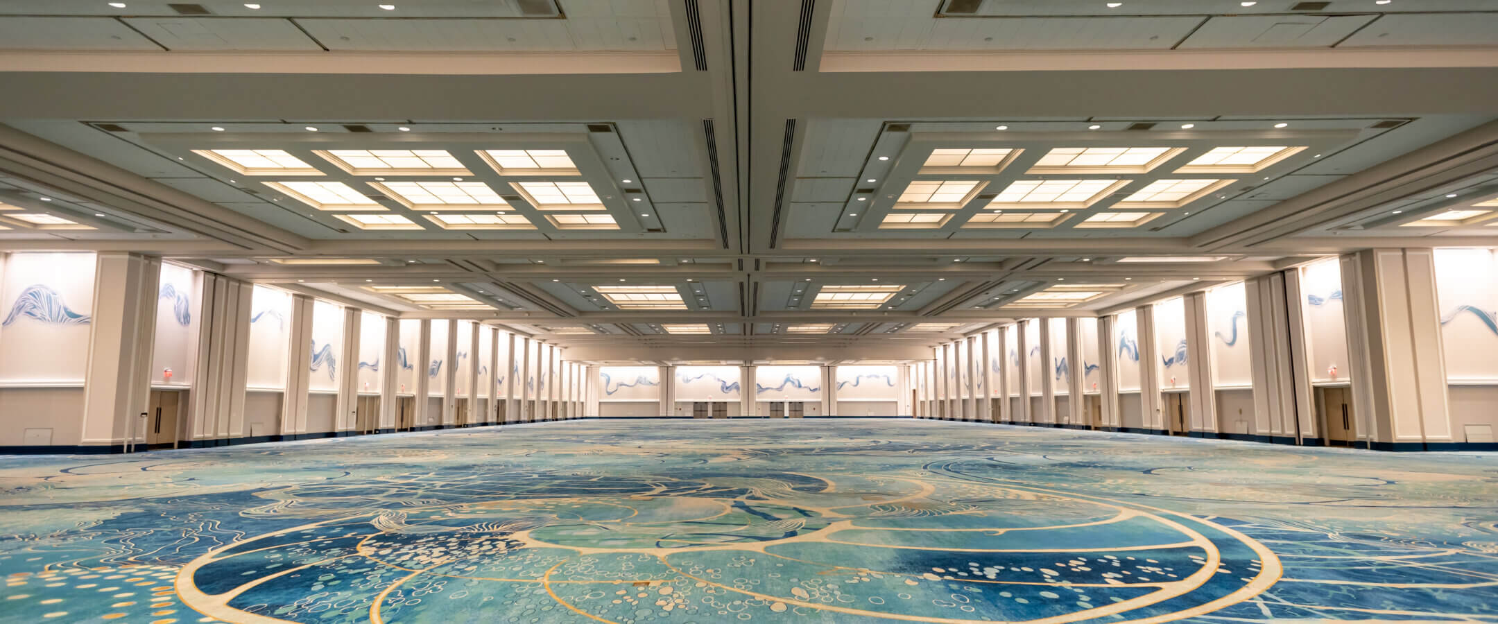 A large, empty convention hall with high ceilings, bright overhead lights, and decorative blue and beige carpeting featuring abstract wave and swirl patterns. White walls are adorned with subtle blue mural designs.