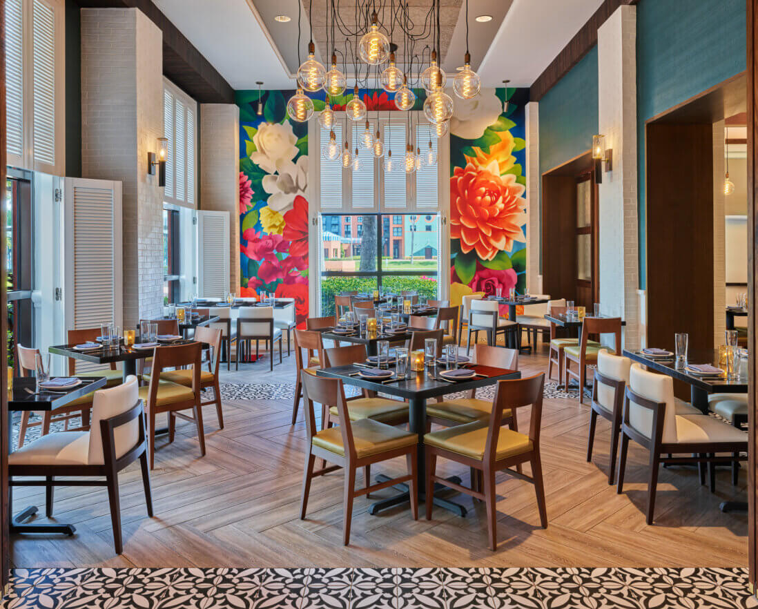 A brightly lit restaurant with wooden tables and chairs, colorful floral murals on the back wall, modern hanging lights, and large windows letting in natural light.