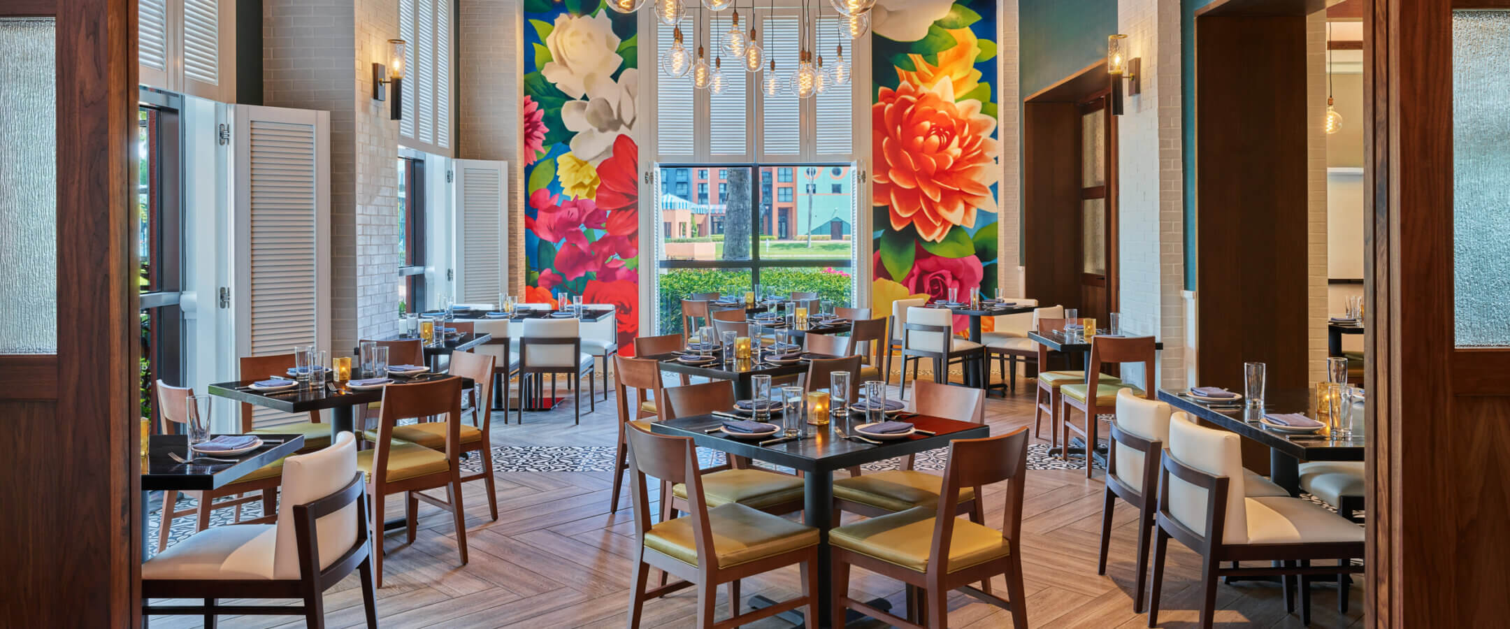 A brightly lit restaurant with wooden tables and chairs, colorful floral murals on the back wall, modern hanging lights, and large windows letting in natural light.