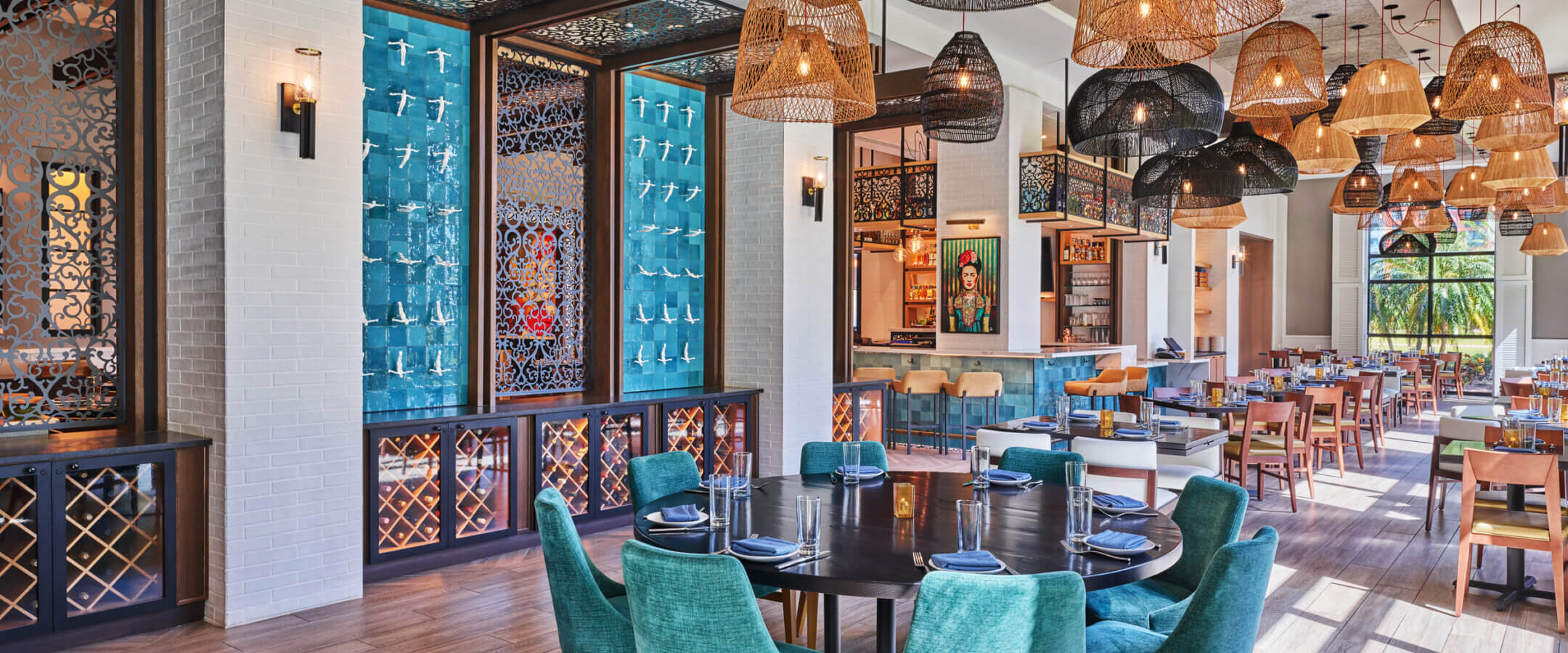 A stylish restaurant interior with teal chairs around round tables, wooden floors, wicker pendant lights, decorative blue wall panels, and large windows letting in natural light.