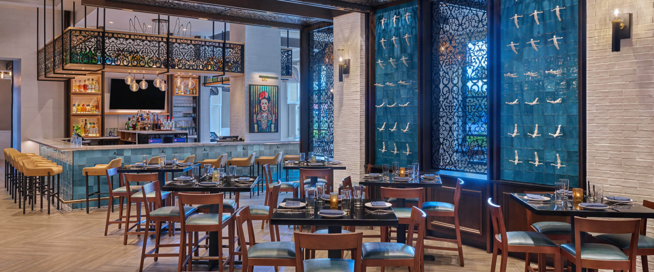 Modern restaurant interior with wooden tables and chairs, a bar area with high stools, elegant lighting, and a decorative blue wall featuring bird sculptures. The atmosphere is stylish and inviting.
