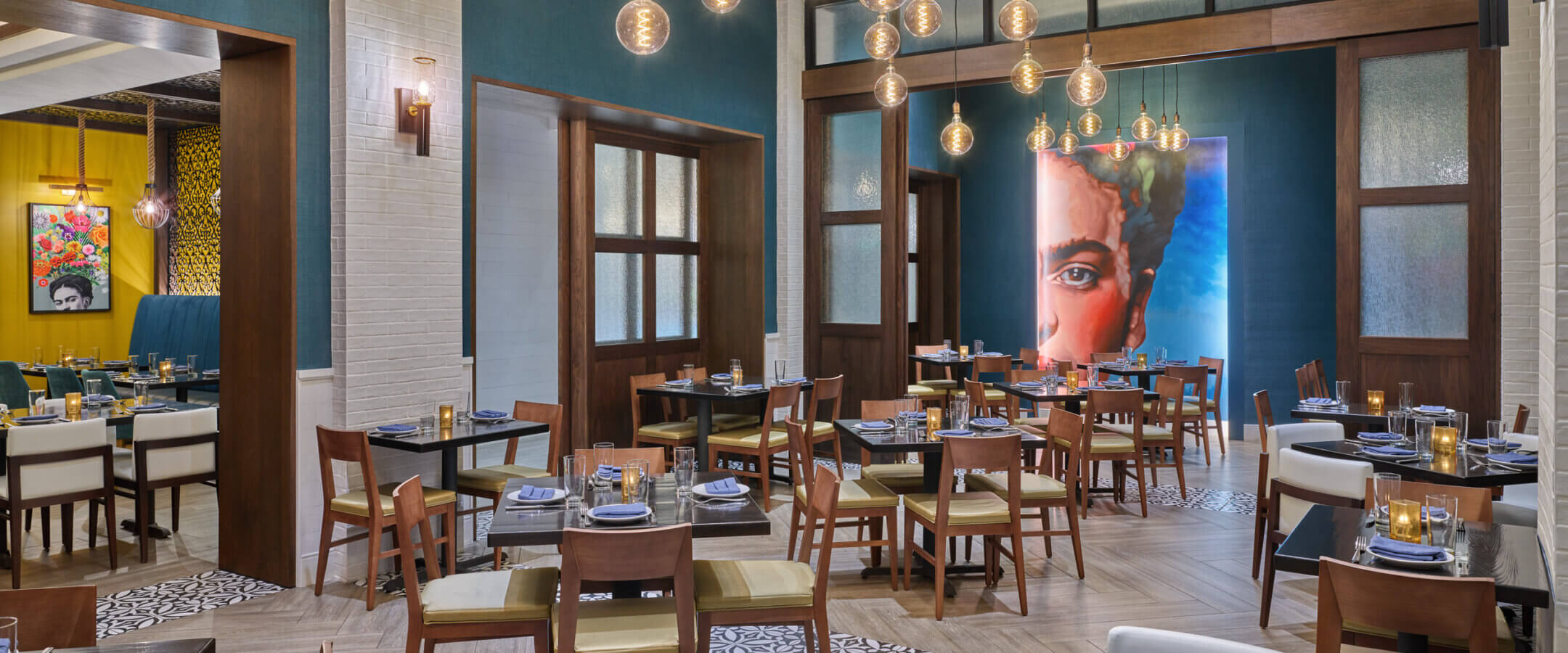 Modern restaurant interior with blue and wood accents, hanging globe lights, neatly set tables, and a large colorful portrait of a womans face on the far wall. Other artwork and seating areas are visible in the background.