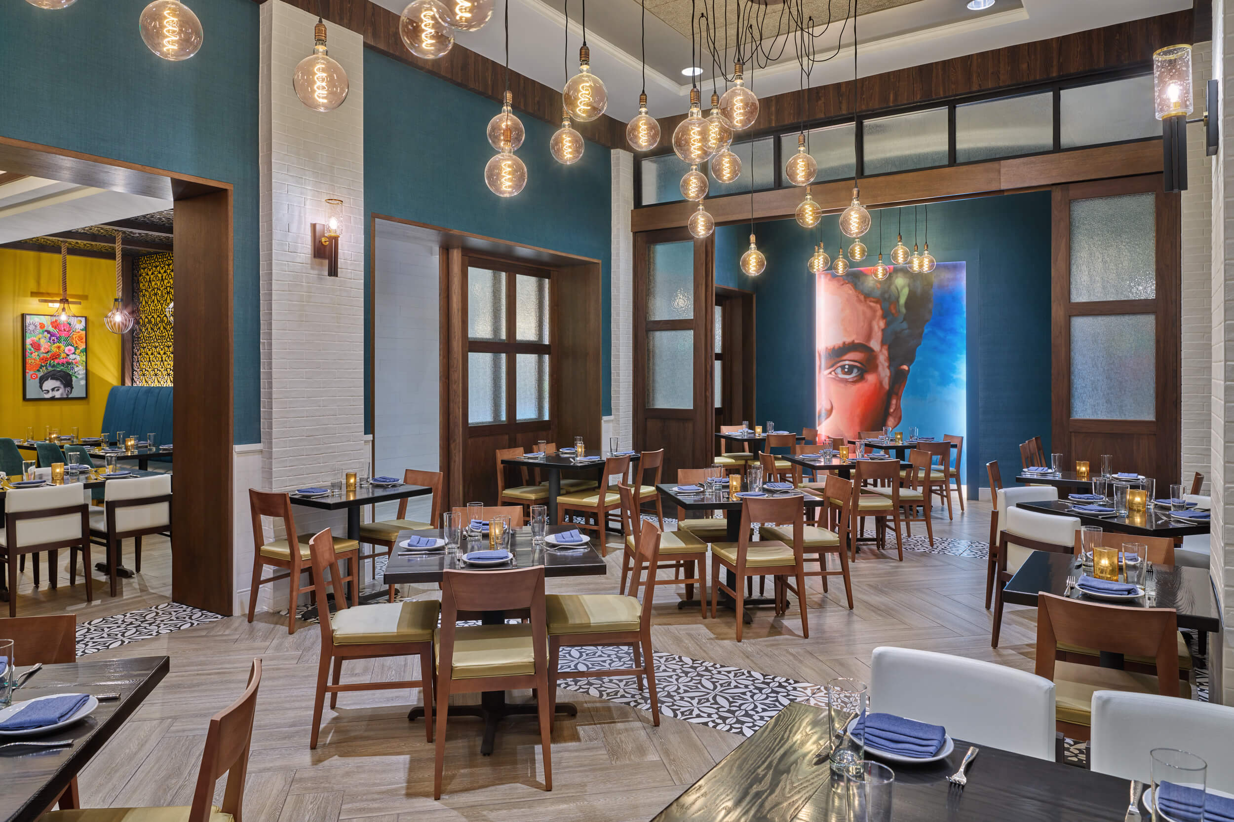 Intérieur d'un restaurant moderne avec des touches de bleu et de bois, des lampes globe suspendues, des tables bien rangées et un grand portrait coloré d'une femme sur le mur du fond. D'autres œuvres d'art et des places assises sont visibles à l'arrière-plan.