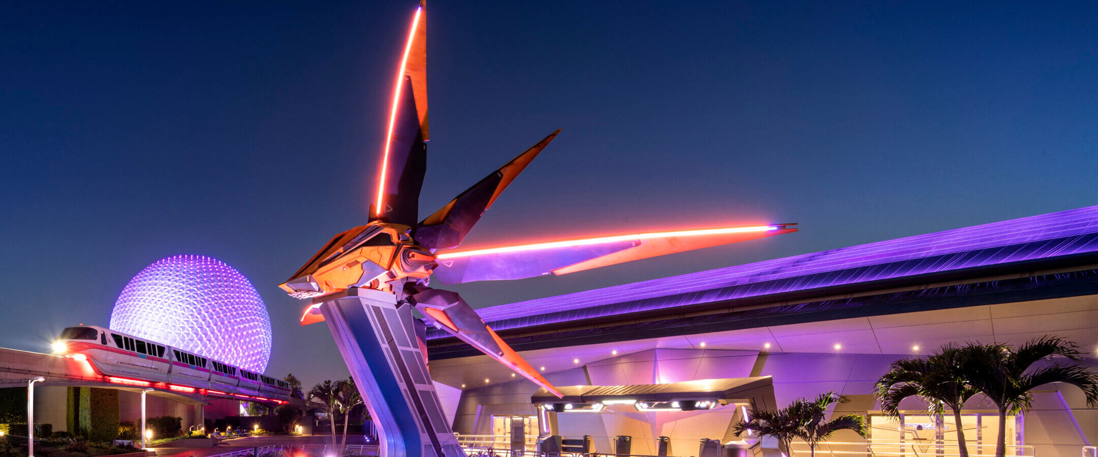 A large, glowing spaceship statue stands illuminated by colorful lights at night, with a futuristic building and the iconic Epcot sphere glowing purple in the background.