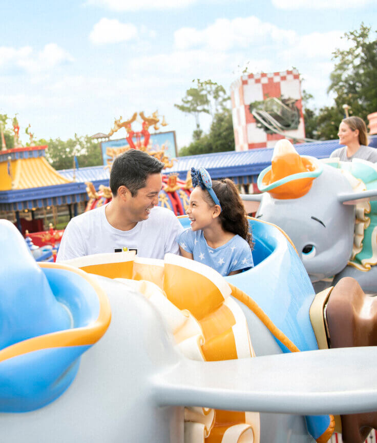A man and a young girl smile at each other while riding a Dumbo-themed amusement park ride, surrounded by colorful decorations and other riders on a sunny day.