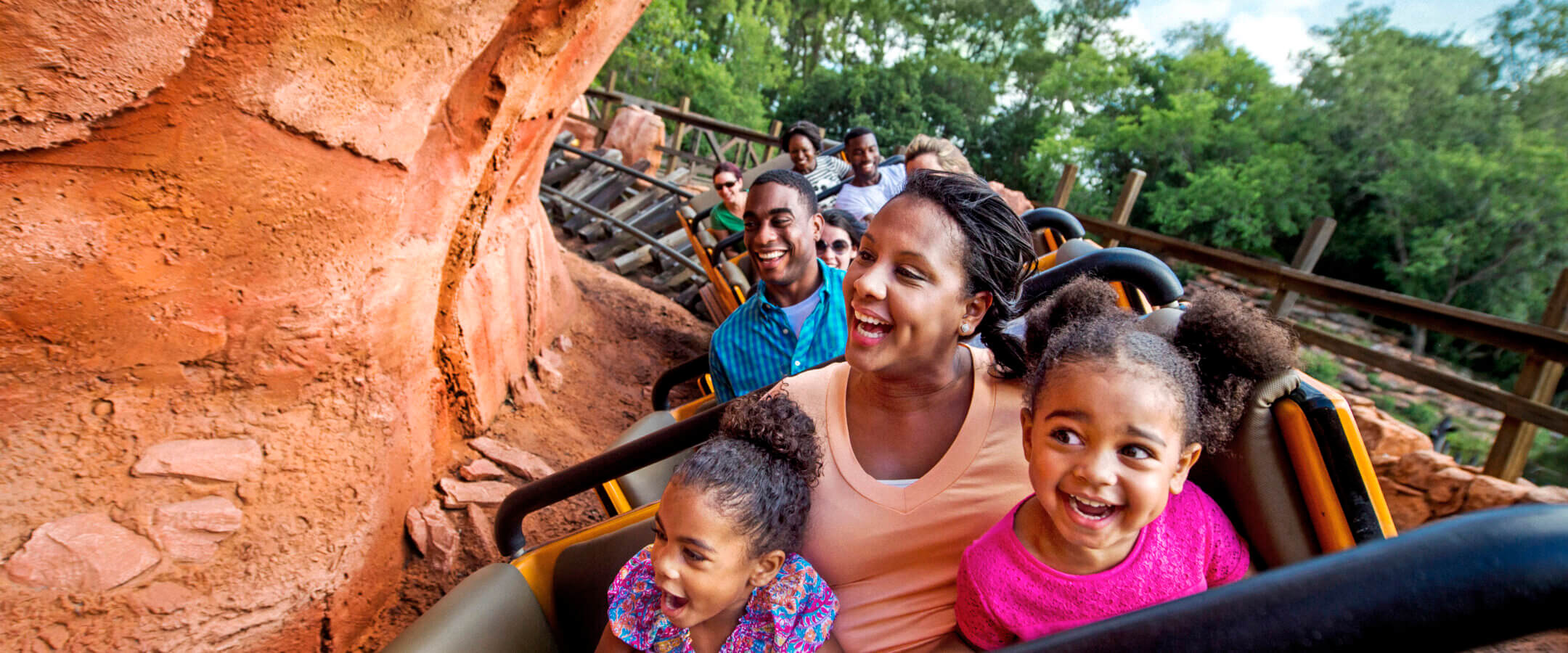 A family rides a roller coaster, smiling and laughing with excitement. The group includes adults and children, surrounded by rocky scenery and green trees under a sunny sky.
