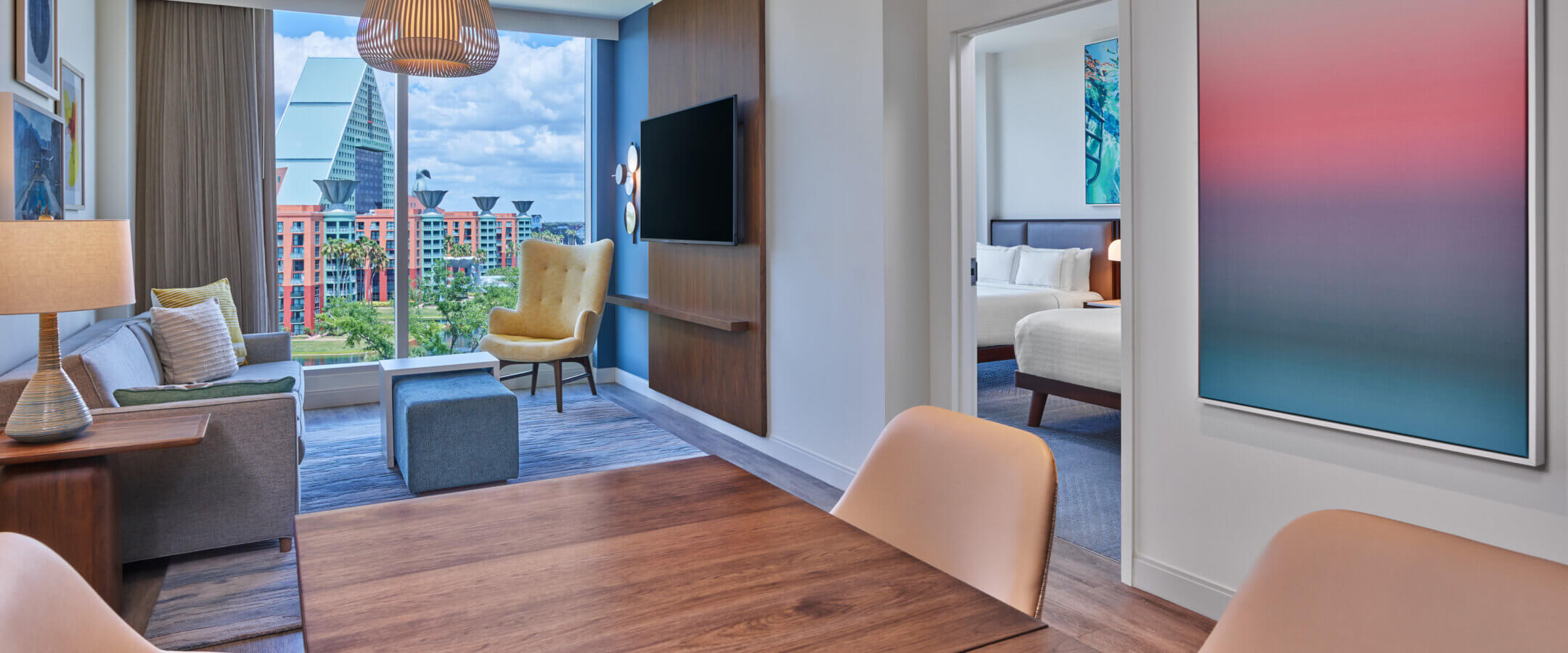 A modern hotel suite with a dining table, beige chairs, a living area with a sofa, large windows showing a cityscape, and a separate bedroom. Contemporary décor features abstract art and wood accents.