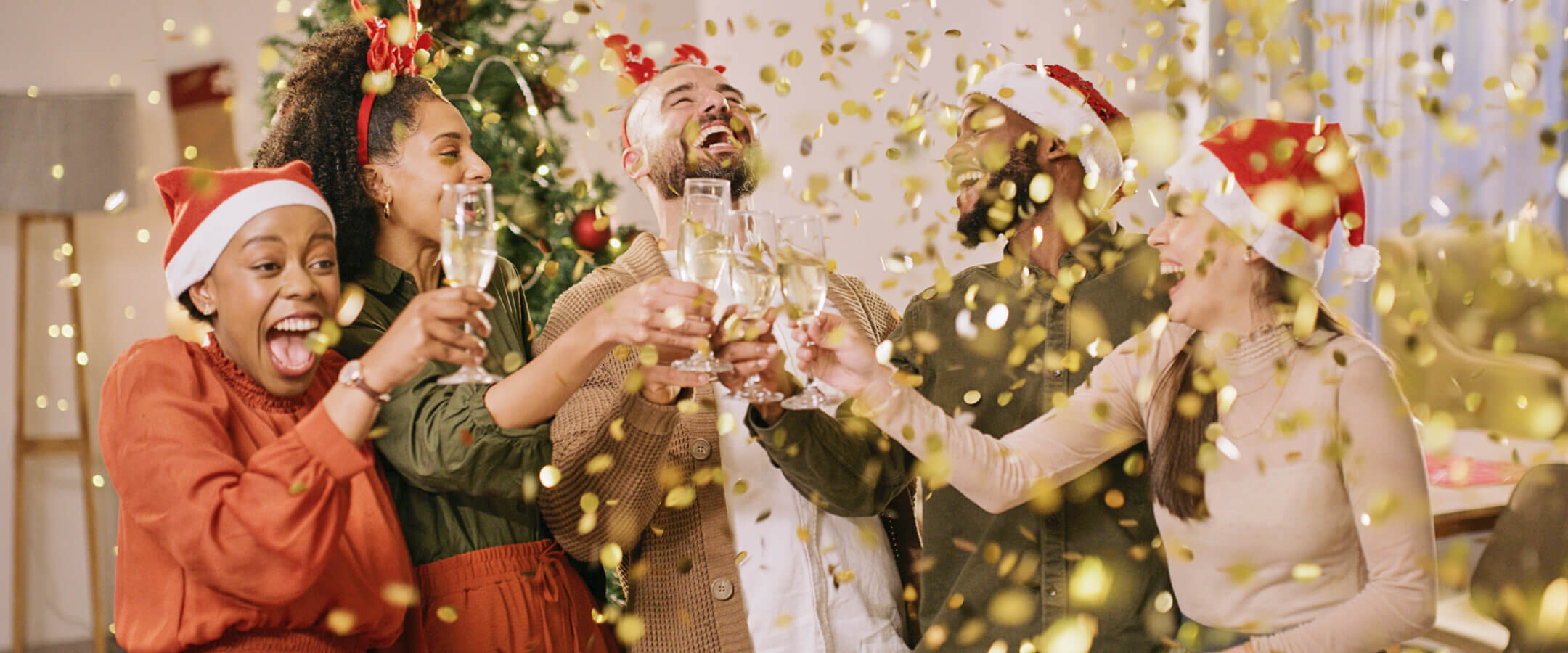 Five people wearing festive outfits and Santa hats clink glasses and celebrate together, surrounded by falling gold confetti, with a decorated Christmas tree in the background.
