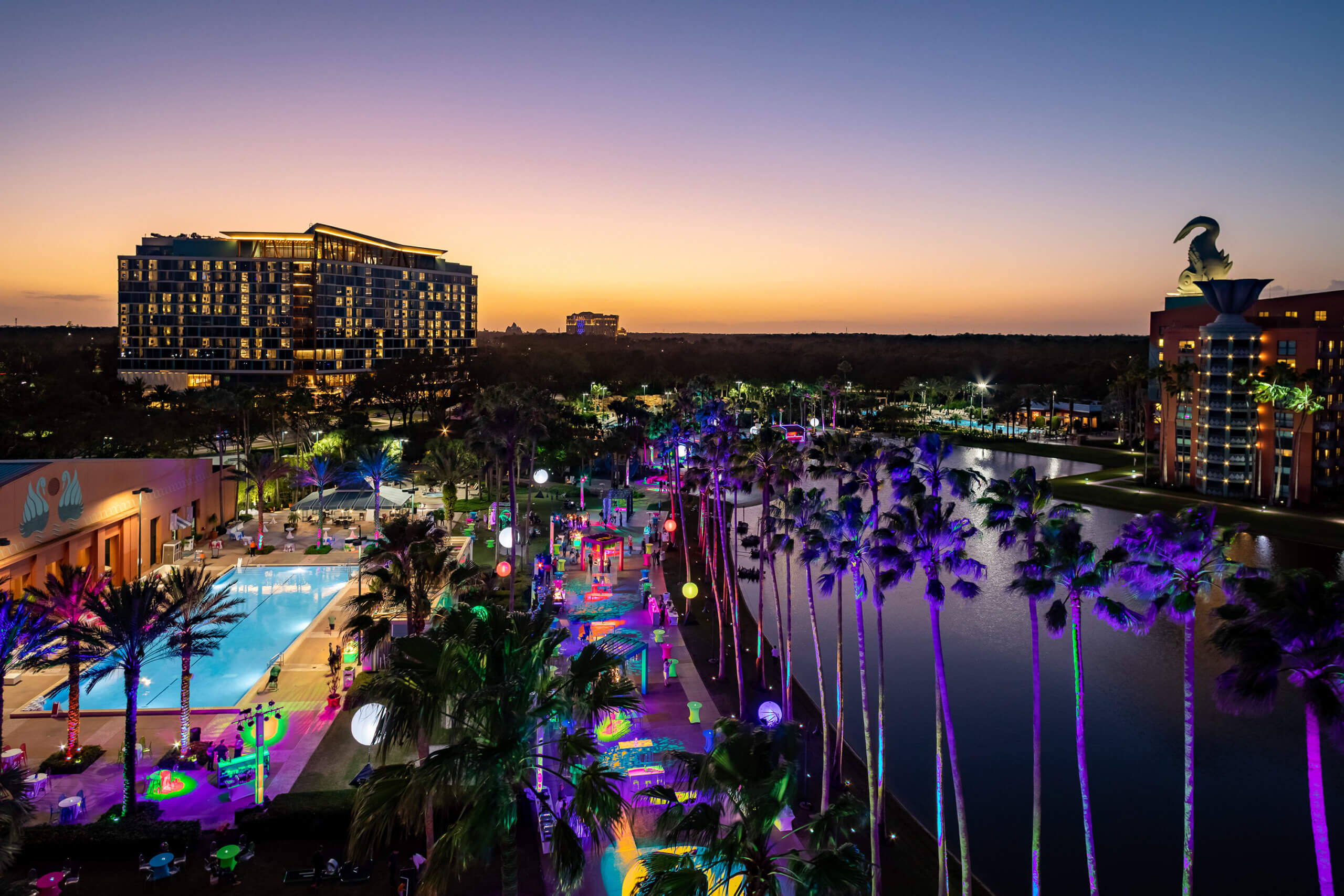A resort at sunset features a brightly lit pool, colorful lights illuminating palm trees and pathways, a large pond, and tall buildings in the background under a clear sky.