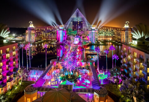 A vibrant nighttime view of a resort with a pyramid-shaped building, dramatic spotlights, and colorful illuminated palm trees and pathways, surrounded by water and modern architecture.