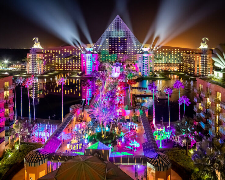 A vibrant nighttime view of a resort with a pyramid-shaped building, dramatic spotlights, and colorful illuminated palm trees and pathways, surrounded by water and modern architecture.