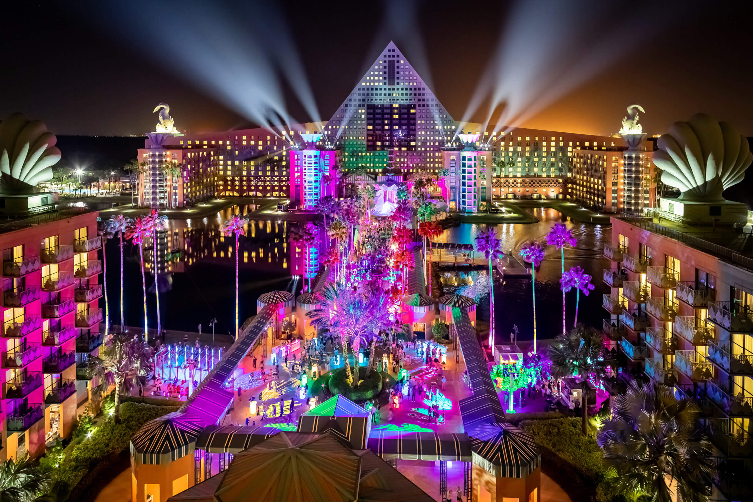 Eine lebendige Nachtansicht eines Resorts mit einem pyramidenförmigen Gebäude, dramatischen Scheinwerfern und farbenfroh beleuchteten Palmen und Wegen, umgeben von Wasser und moderner Architektur.