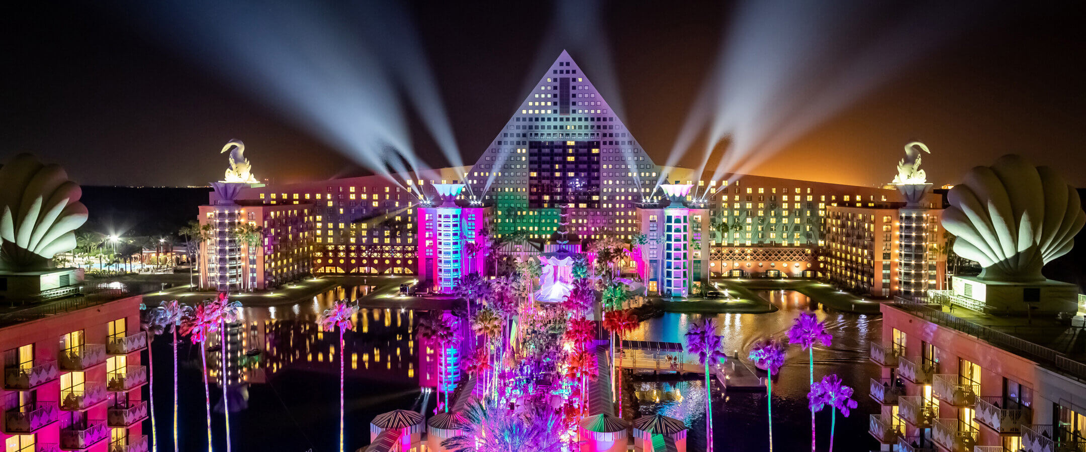 A brightly lit hotel with a pyramid-shaped center and statues on each side glows at night, with palm trees and colorful lights illuminating the foreground and beams shining skyward.