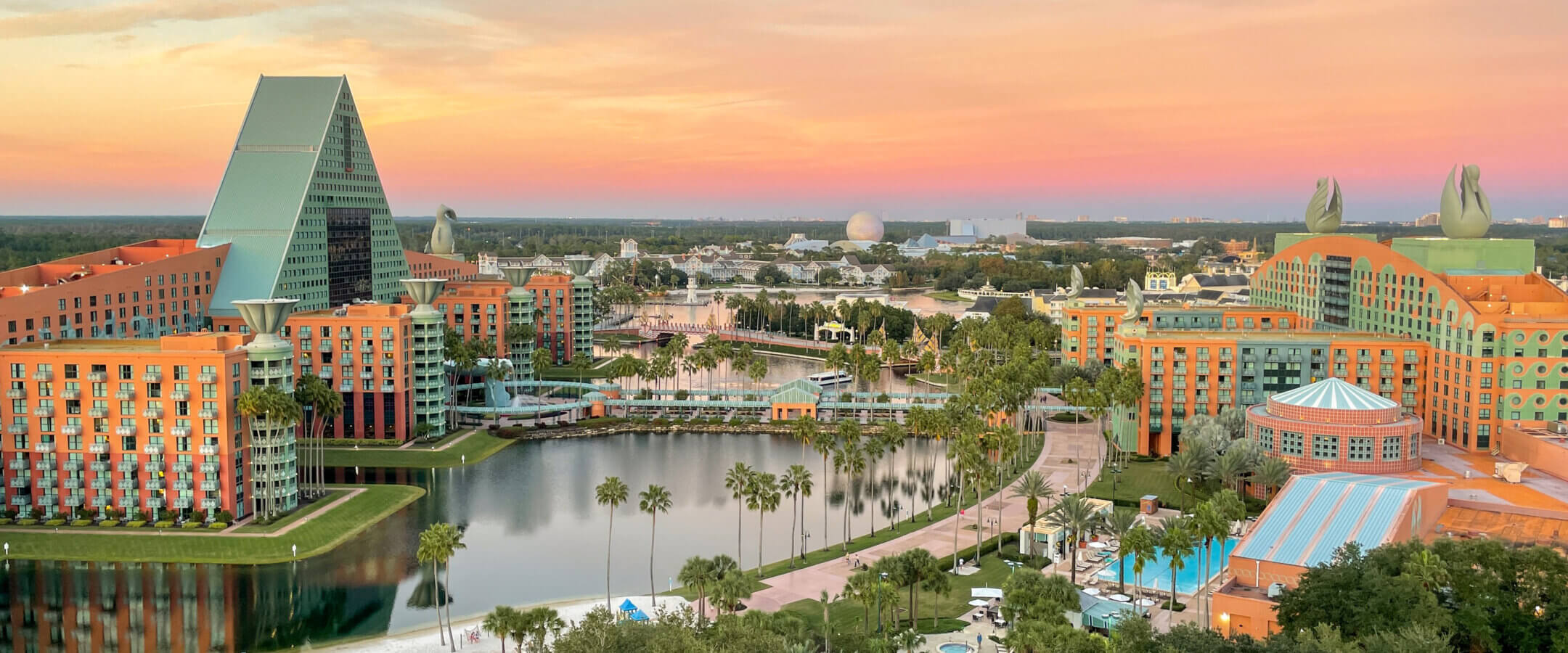A scenic view of colorful resort buildings with unique triangular and swan-shaped rooftops, a calm lake, palm trees, and a sunset sky with pink and orange hues in the background.