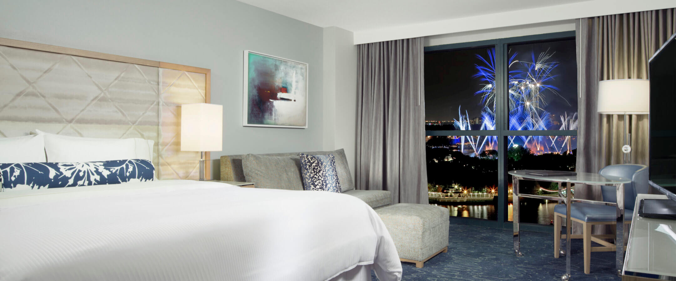 A modern hotel room with a large bed, a sofa, a desk, and a window showing a cityscape at night with colorful fireworks lighting up the sky.