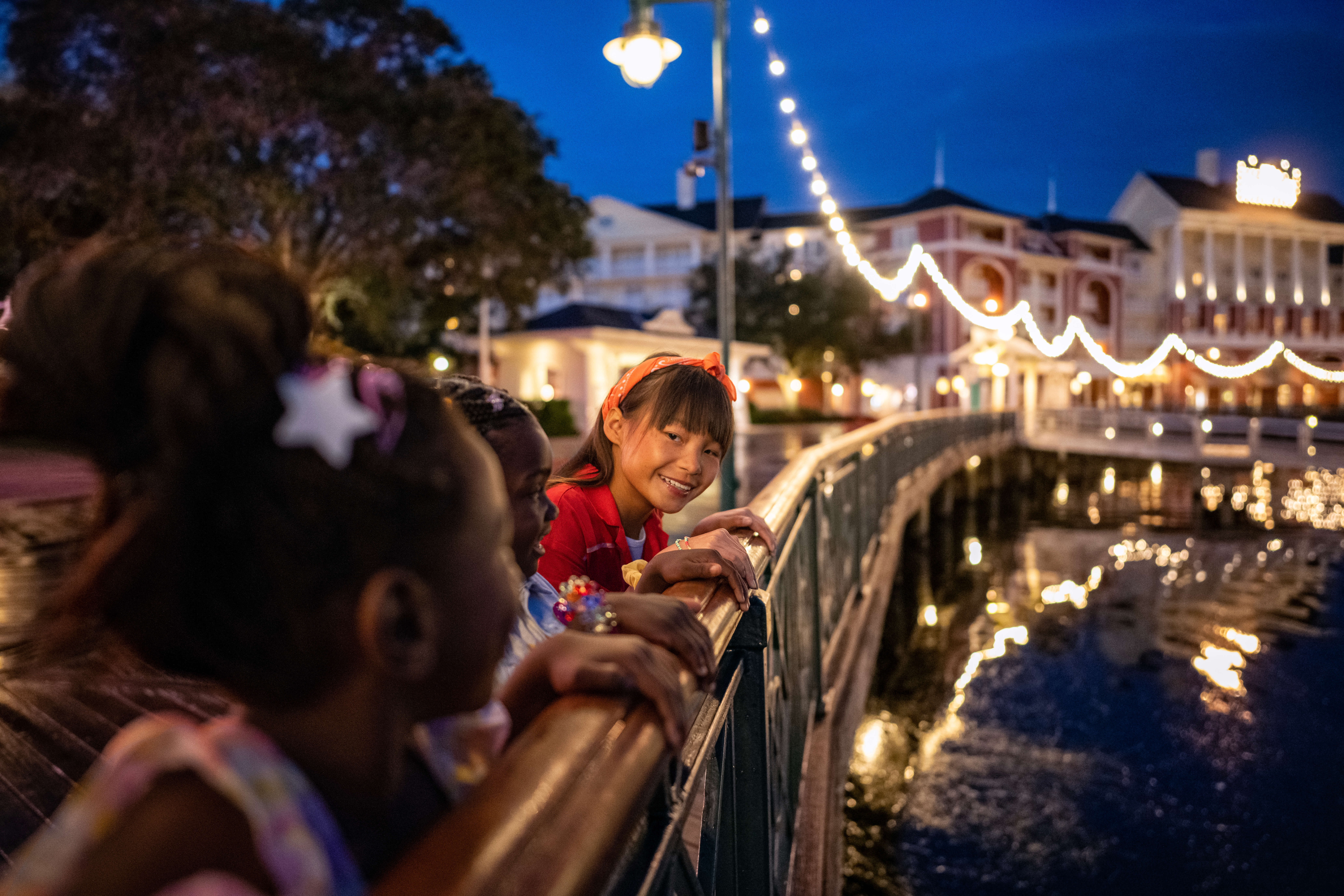 Three children stand on a boardwalk at night, looking over a railing at water. String lights and buildings are visible in the background, creating a festive and warm atmosphere.